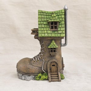 May include: A whimsical fairy house shaped like a brown boot with a green roof and chimney. The boot has a wooden door, windows, and a small porch. The boot is decorated with faux laces and sits on a stone base with green foliage.