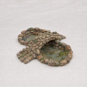 May include: A miniature stone bridge with two ponds. The bridge is made of small, textured stones in shades of brown and gray, with patches of green moss. The ponds have a water-like appearance.