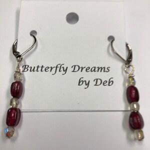 May include: A pair of dangle earrings with silver-tone hooks. Each earring features a series of beads: three maroon, two clear, and one iridescent. The earrings are displayed on a white card with the text "Butterfly Dreams by Deb."