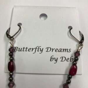 May include: Dangling earrings with a dark red, oval-shaped bead, dark gray beads, and purple crystal accents. The earrings are attached to silver-colored lever-back ear wires. The earrings are displayed on a white card with the text "Butterfly Dreams by Deb".