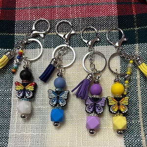 May include: Four keychains with butterfly charms, each featuring a different color scheme. The keychains have silver rings and clasps, with beaded accents and tassels in yellow, blue, and purple. The butterfly charms are decorated with colorful details.