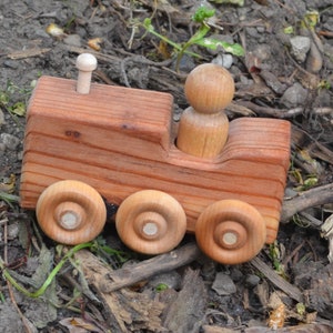 May include: A wooden toy tractor with a small wooden figure in the driver's seat. The tractor has four wheels and a chimney on top. The toy is made of natural wood and has a rustic finish.