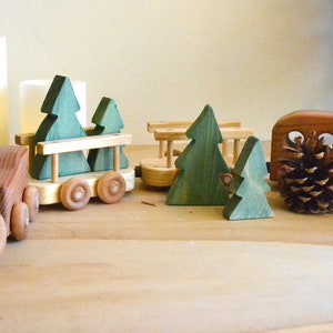 May include: A wooden toy train set with a brown engine, two boxcars, and a flatbed car carrying three green wooden trees. The train is on a wooden surface.