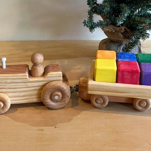 May include: A wooden toy tractor with a trailer hauling a stack of colorful wooden blocks. The tractor is made of light wood with brown stripes and has a small wooden figure in the driver's seat. The trailer is filled with six blocks in shades of orange, yellow, red, green, and purple.
