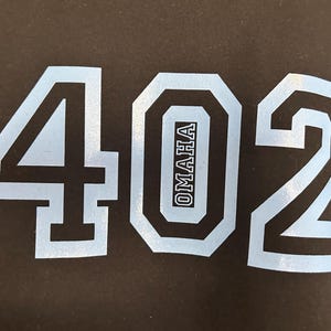 May include: Black fabric with the light blue numbers "402" in a bold, blocky font. The number "0" has the word "OMAHA" in a vertical, light blue font inside.