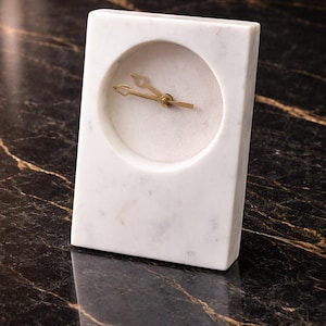 May include: A rectangular white marble clock with a circular clock face. The clock hands are gold-colored. The clock is set on a black marble surface with gold veining.