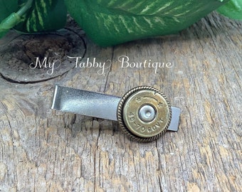 May include: A silver-toned metal tie clip featuring a circular brass 45-70 GOVT cartridge head casing with a rope-textured border. The text My Tabby Boutique is visible in the upper portion of the image above the tie clip.