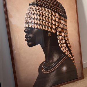 May include: A framed wall art print depicting a profile view of a person with dark skin wearing a headpiece covered in numerous cream-colored cowrie shells that partially obscure the eyes. The person wears a simple gold stud earring and a layered necklace.