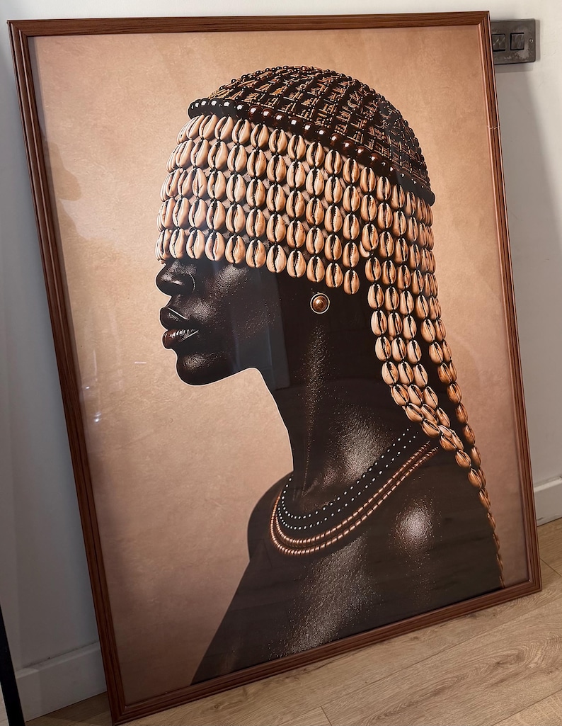 May include: A framed wall art print depicting a profile view of a person with dark skin wearing a headpiece covered in numerous cream-colored cowrie shells that partially obscure the eyes. The person wears a simple gold stud earring and a layered necklace.