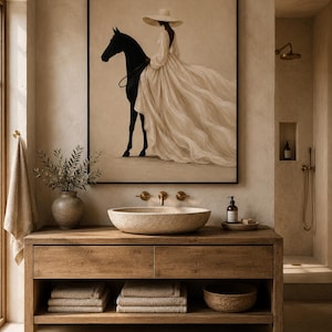 May include: A framed wall art print featuring a figure in a wide-brimmed hat and a flowing cream-colored gown riding a black horse. The artwork is displayed above a wooden bathroom vanity with a stone vessel sink and stacked beige towels.