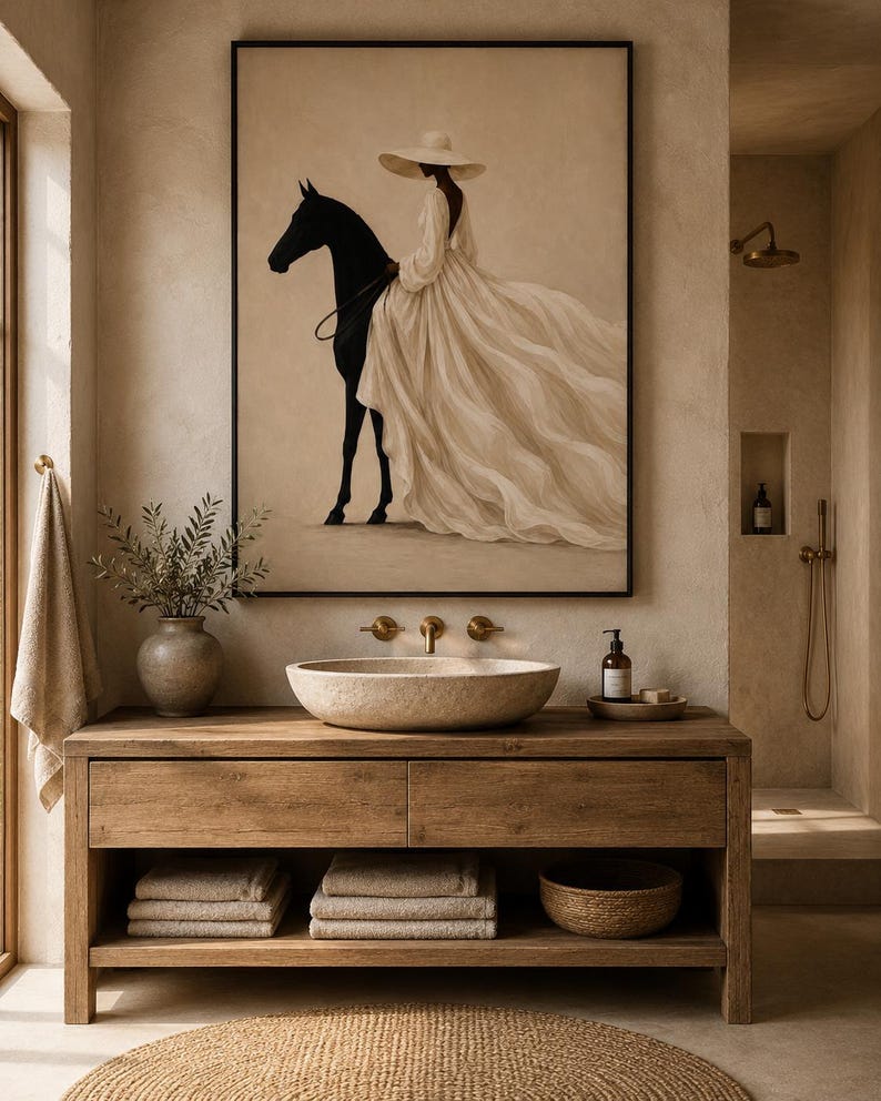 May include: A framed wall art print featuring a figure in a wide-brimmed hat and a flowing cream-colored gown riding a black horse. The artwork is displayed above a wooden bathroom vanity with a stone vessel sink and stacked beige towels.