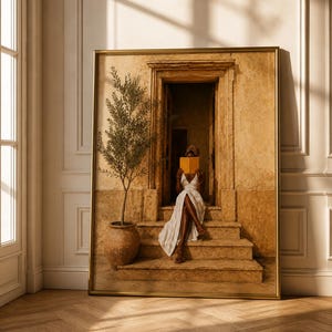 May include: A framed wall art print depicting a person in a white dress sitting on stone steps in a doorway, holding an open yellow book. Beside the person is a potted olive tree in a terracotta vessel, all rendered in warm, golden-brown tones.