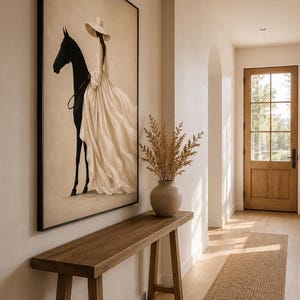 May include: A framed rectangular wall art piece featuring a painting of a person wearing a wide-brimmed hat and a long, flowing cream-colored dress, standing beside a solid black horse against a neutral, light beige background.