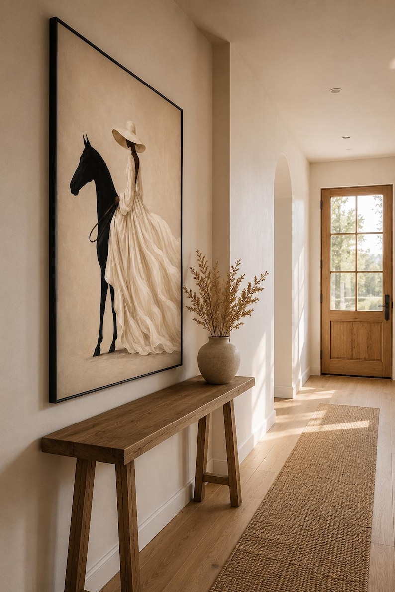 May include: A framed rectangular wall art piece featuring a painting of a person wearing a wide-brimmed hat and a long, flowing cream-colored dress, standing beside a solid black horse against a neutral, light beige background.