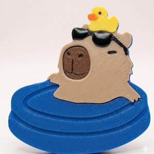 May include: A whimsical 3D-printed capybara figurine. The capybara is tan with a brown face, wearing a black eye patch and a yellow rubber ducky on its head. It appears to be floating in a blue pool. A fun, decorative item.