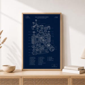 May include: A framed wall art print featuring a dark blue background with a white technical patent diagram of a 35mm rangefinder camera. The print includes detailed exploded view illustrations, numbered parts, and descriptive text labels in English.
