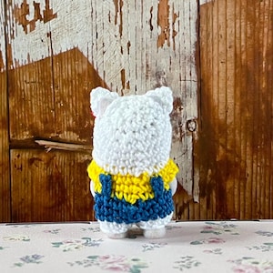 Puede incluir: Figura de oso de ganchillo blanco hecha a mano con una camisa amarilla y un peto azul. El oso tiene peque&ntilde;as orejas blancas y est&aacute; de pie sobre una superficie con estampado floral. El fondo es una pared de madera r&uacute;stica.