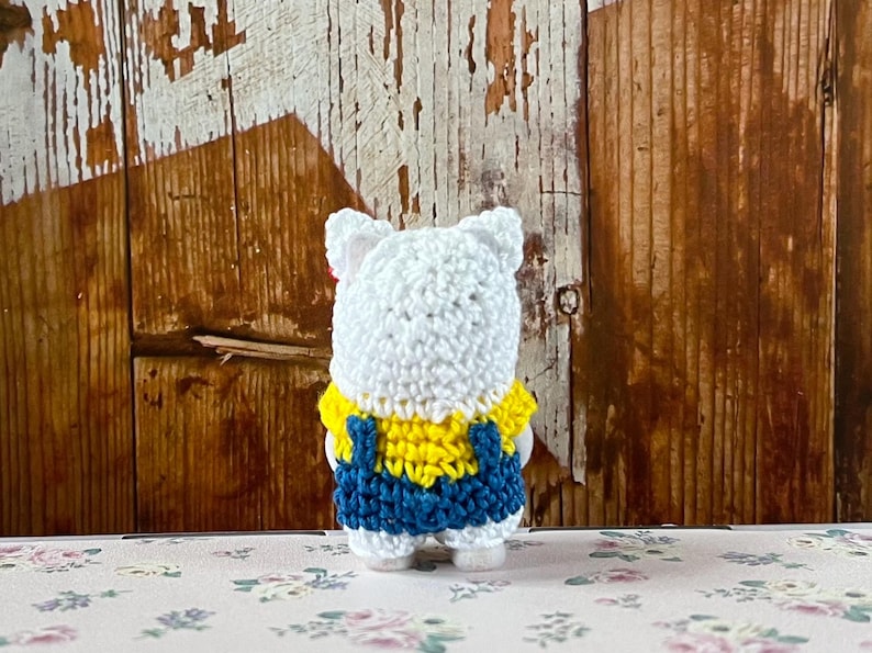 Puede incluir: Figura de oso de ganchillo blanco hecha a mano con una camisa amarilla y un peto azul. El oso tiene peque&ntilde;as orejas blancas y est&aacute; de pie sobre una superficie con estampado floral. El fondo es una pared de madera r&uacute;stica.