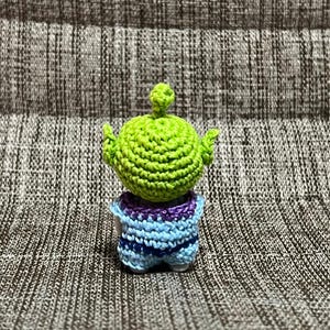 May include: A handmade crochet alien toy in green, blue, and purple. The alien has a large green head with small arms raised. The body is blue and purple. The toy is sitting on a textured gray surface.