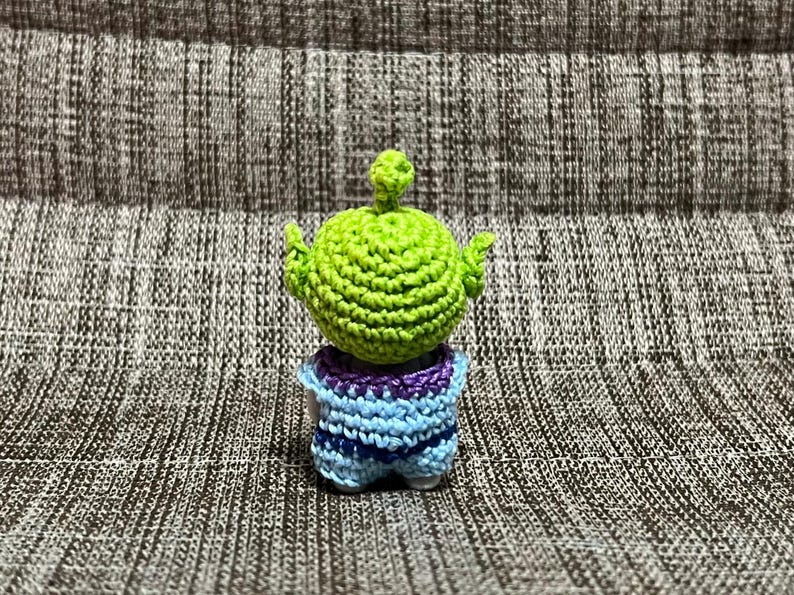 May include: A handmade crochet alien toy in green, blue, and purple. The alien has a large green head with small arms raised. The body is blue and purple. The toy is sitting on a textured gray surface.