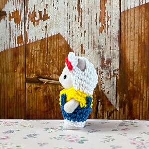 Puede incluir: Peque&ntilde;a figura de conejo blanco con un atuendo de ganchillo. El atuendo incluye un sombrero blanco con una banda roja, un cuello amarillo y un chaleco azul. El fondo es una pared de madera r&uacute;stica.
