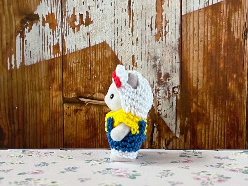 Puede incluir: Peque&ntilde;a figura de conejo blanco con un atuendo de ganchillo. El atuendo incluye un sombrero blanco con una banda roja, un cuello amarillo y un chaleco azul. El fondo es una pared de madera r&uacute;stica.