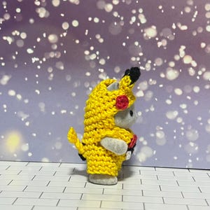 Puede incluir: Una peque&ntilde;a figura gris con un disfraz de Pikachu amarillo tejido a mano. El disfraz incluye una capucha con orejas negras y un detalle de mejilla roja. La figura est&aacute; sobre una superficie de ladrillo blanco con un fondo bokeh morado y blanco.