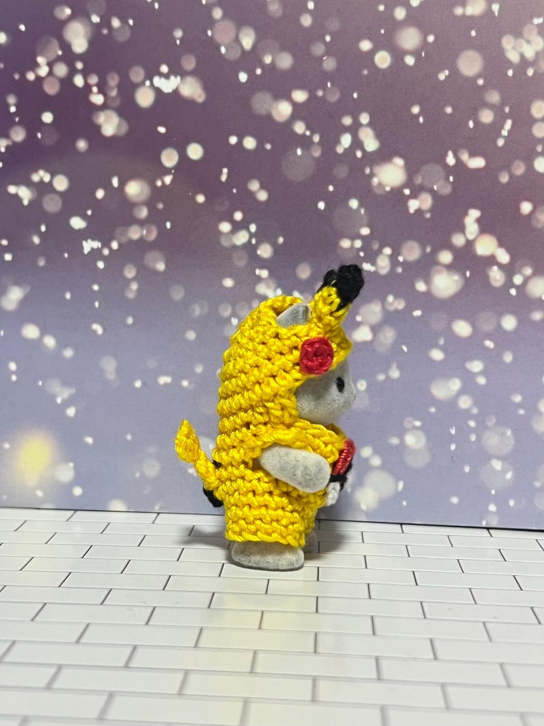 Puede incluir: Una peque&ntilde;a figura gris con un disfraz de Pikachu amarillo tejido a mano. El disfraz incluye una capucha con orejas negras y un detalle de mejilla roja. La figura est&aacute; sobre una superficie de ladrillo blanco con un fondo bokeh morado y blanco.