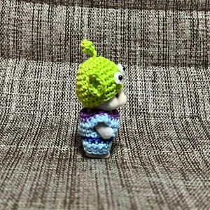 May include: A small, handmade figurine of a green alien character, wearing a lime green crocheted hat and a blue and purple striped crocheted shirt. The alien has large, googly eyes.