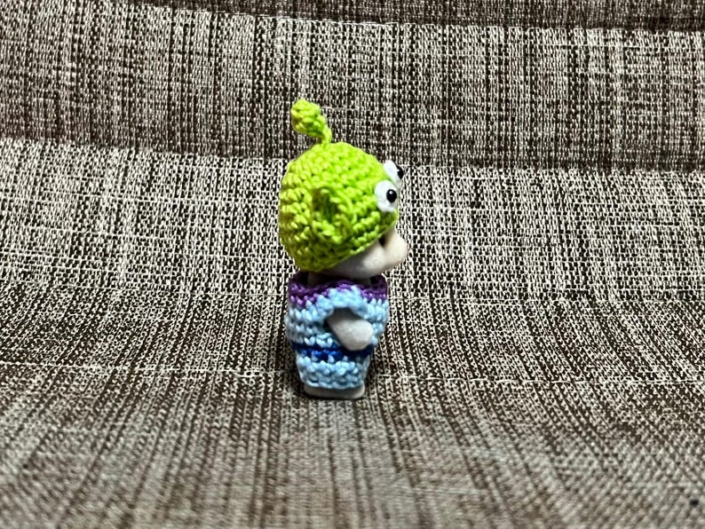 May include: A small, handmade figurine of a green alien character, wearing a lime green crocheted hat and a blue and purple striped crocheted shirt. The alien has large, googly eyes.