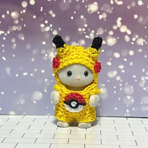 Puede incluir: Una peque&ntilde;a figura blanca con un disfraz de Pikachu amarillo tejido a mano, con orejas negras, detalles rojos y un emblema de Pok&eacute;ball. El fondo es un bokeh de luces moradas y blancas.