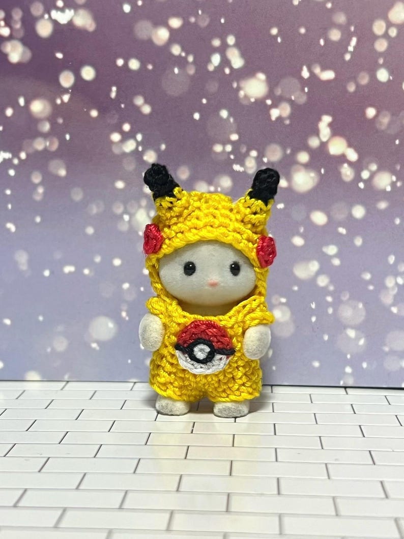 Puede incluir: Una peque&ntilde;a figura blanca con un disfraz de Pikachu amarillo tejido a mano, con orejas negras, detalles rojos y un emblema de Pok&eacute;ball. El fondo es un bokeh de luces moradas y blancas.