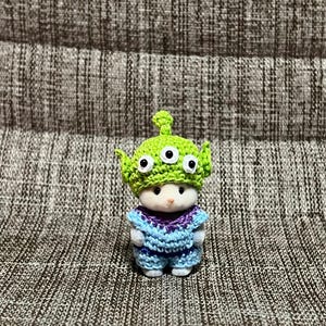 May include: A small, white figurine wearing a crocheted outfit and a green alien hat with three eyes. The outfit is blue and purple, with a textured pattern. The figurine is standing on a textured surface.