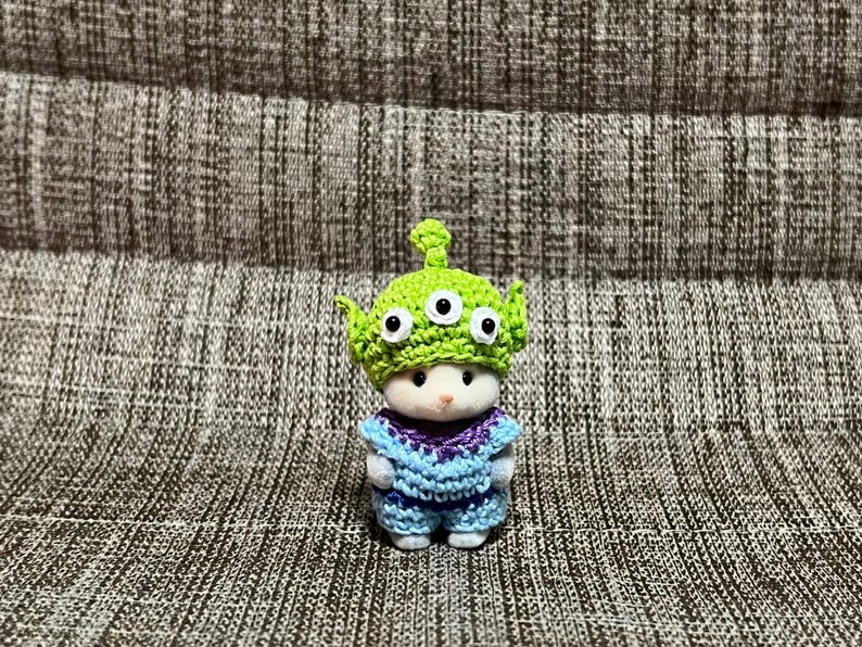 May include: A small, white figurine wearing a crocheted outfit and a green alien hat with three eyes. The outfit is blue and purple, with a textured pattern. The figurine is standing on a textured surface.