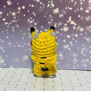 Puede incluir: Una figura de Pikachu de ganchillo amarillo hecha a mano, vista desde atr&aacute;s. La figura tiene orejas con puntas negras, orejas internas grises y una boca roja. Se ven rayas negras en la espalda. La figura est&aacute; sobre una superficie de baldosas blancas y grises.