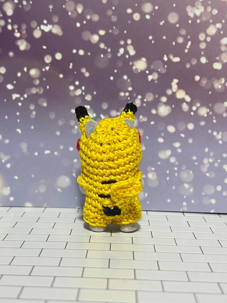 Puede incluir: Una figura de Pikachu de ganchillo amarillo hecha a mano, vista desde atr&aacute;s. La figura tiene orejas con puntas negras, orejas internas grises y una boca roja. Se ven rayas negras en la espalda. La figura est&aacute; sobre una superficie de baldosas blancas y grises.