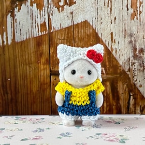 Puede incluir: Una peque&ntilde;a figura blanca con un atuendo hecho a mano. El atuendo incluye un gorro de ganchillo blanco con un lazo rojo, una parte superior amarilla y un peto azul. La figura est&aacute; sobre una superficie con estampado floral.