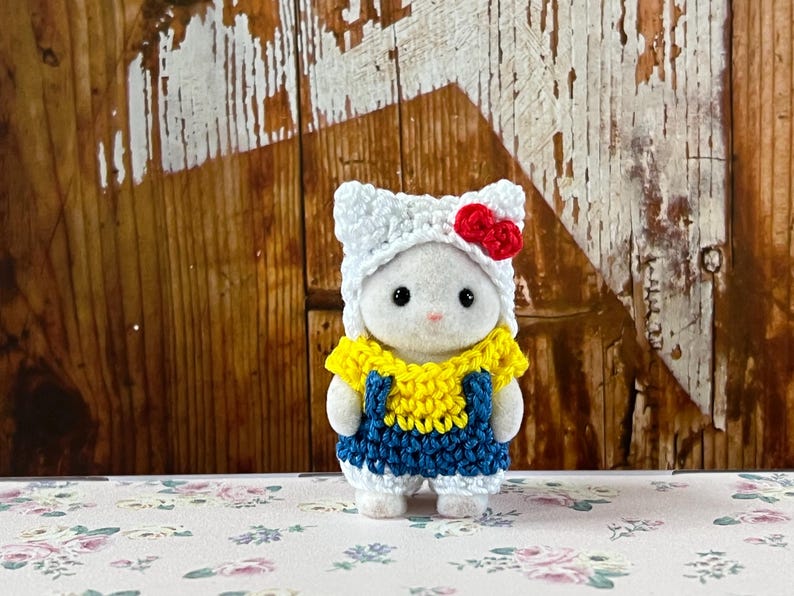 Puede incluir: Una peque&ntilde;a figura blanca con un atuendo hecho a mano. El atuendo incluye un gorro de ganchillo blanco con un lazo rojo, una parte superior amarilla y un peto azul. La figura est&aacute; sobre una superficie con estampado floral.