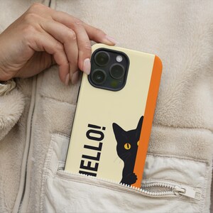 May include: A phone case with a cream-colored background, featuring the word "HELLO!" in black and a black cat peeking from the side. The case has an orange stripe and is in a pocket.