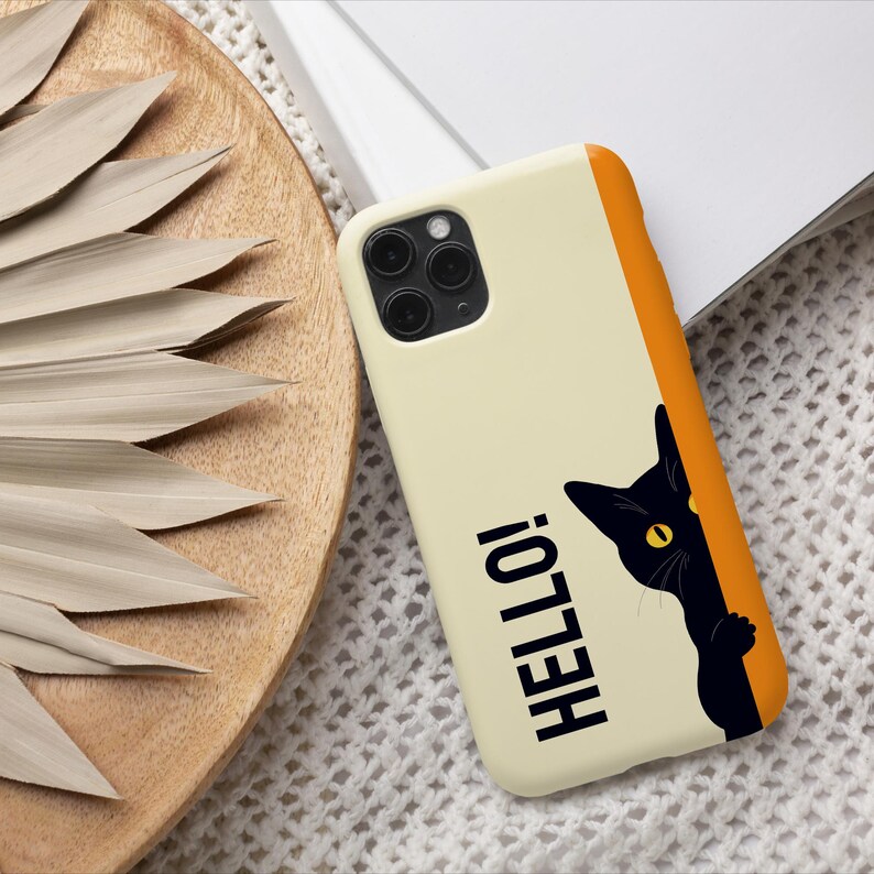 May include: A cream-colored phone case with an orange border and the word "HELLO!" in black. A black cat peeks from the side with yellow eyes. The case is on a white surface with a wooden tray and dried leaves.