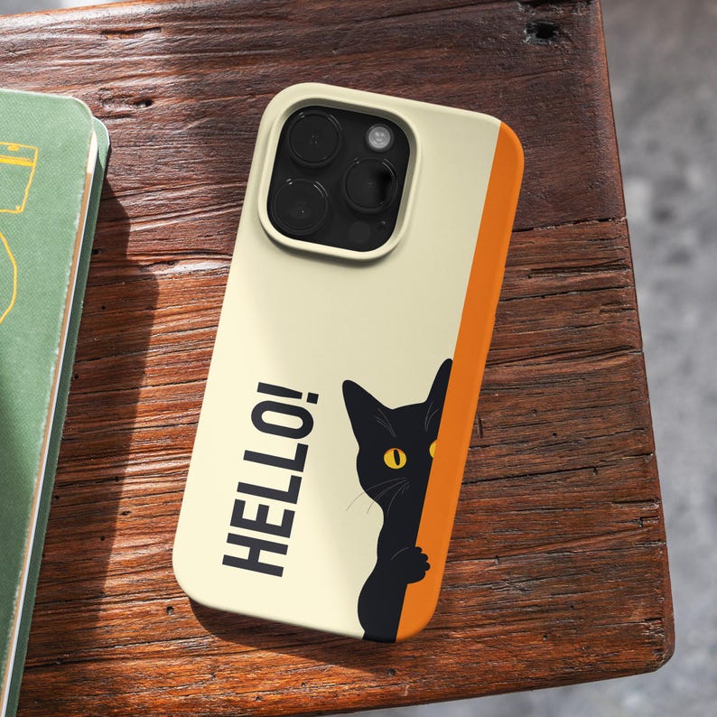 May include: A phone case with a cream-colored background and an orange stripe on the side. The case features a black cat peeking out with a yellow eye and the word "HELLO!" in black.