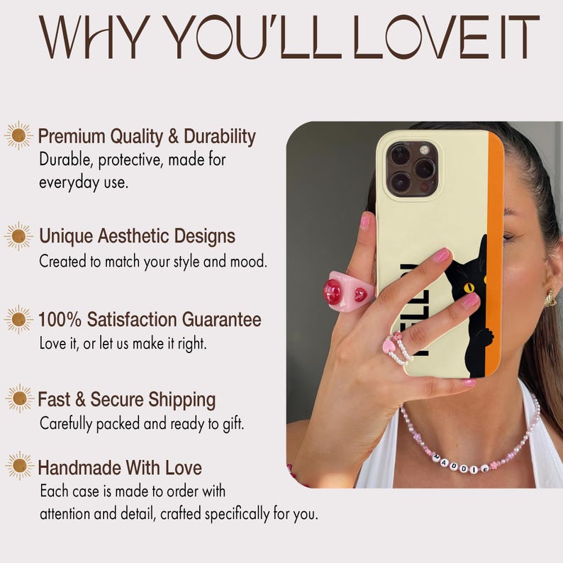 May include: A phone case with a black cat design and the word "MADDI" in black. The case has a cream-colored background with an orange stripe. The image also shows a person wearing rings and a necklace.