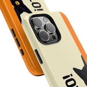 May include: Cream-colored phone case with a black cat illustration and the word "!o". The case has a black border around the camera lenses and a bright orange border. The phone case is designed to protect the phone from scratches and damage.