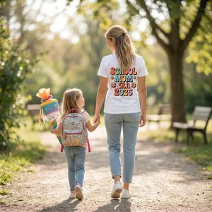 Könnte beinhalten: Ein weißes T-Shirt mit dem bunten Schriftzug SCHOOL MOM ERA 2026 auf der Rückseite, kombiniert mit einer blauen Jeans. Ein Kind trägt einen Rucksack mit Blumenmuster und eine bunte, kegelförmige Geschenktüte mit Bändern, ebenfalls in hellem Oberteil.