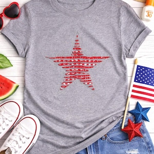 May include: A gray t-shirt with a distressed red star design. The image also includes denim shorts, white sneakers, sunglasses, a straw hat, and patriotic decorations, suggesting a theme of American pride and summer.