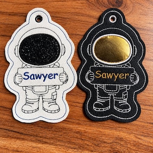 Puede incluir: Dos llaveros con forma de astronauta y contornos bordados. El llavero izquierdo es blanco con una visera de purpurina negra y el texto azul Sawyer. El derecho es negro con una visera dorada y el texto naranja Sawyer.