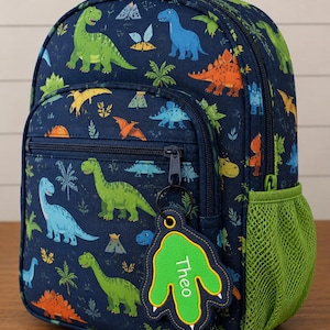 May include: A navy blue backpack featuring a colorful dinosaur pattern with green, blue, and orange prehistoric creatures. The bag includes a lime green mesh side pocket, a top handle, and a hanging tag shaped like a dinosaur footprint with the name Theo.