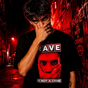 Könnte beinhalten: Schwarzes T-Shirt mit dem Wort "RAVE" in einem weißen und roten Feld und einer Grafik eines Gesichts mit einer Hand über dem Mund. Der Text "IS NOT A CRIME" steht darunter. Der Hintergrund ist eine rot getönte Gasse mit Graffiti.