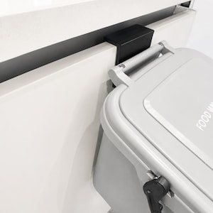 May include: A light grey rectangular waste bin with a hinged lid featuring the text FOOD WASTE. The bin includes a black plastic mounting bracket that hooks over the top edge of a white cabinet door, allowing the container to hang securely.