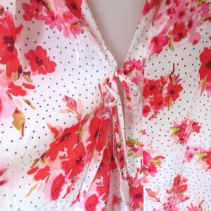 May include: A white blouse with a floral pattern of red and pink flowers. The blouse has a polka dot design and a tie closure at the neckline. The fabric appears to be lightweight and flowy.